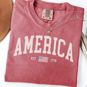 Peut inclure: Un t-shirt rouge avec le mot "AMERICA" en grandes lettres blanches. En dessous du mot "AMERICA" se trouve le texte "EST 1776" et un petit drapeau am&eacute;ricain. Un collier en or avec un pendentif est sur le t-shirt.