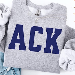 May include: A heather gray sweatshirt with the word "ACK" in navy blue block letters. The sweatshirt has a crew neck and long sleeves with rolled-up cuffs. A silver necklace with a small pendant is draped across the top. A pair of blue jeans is visible.