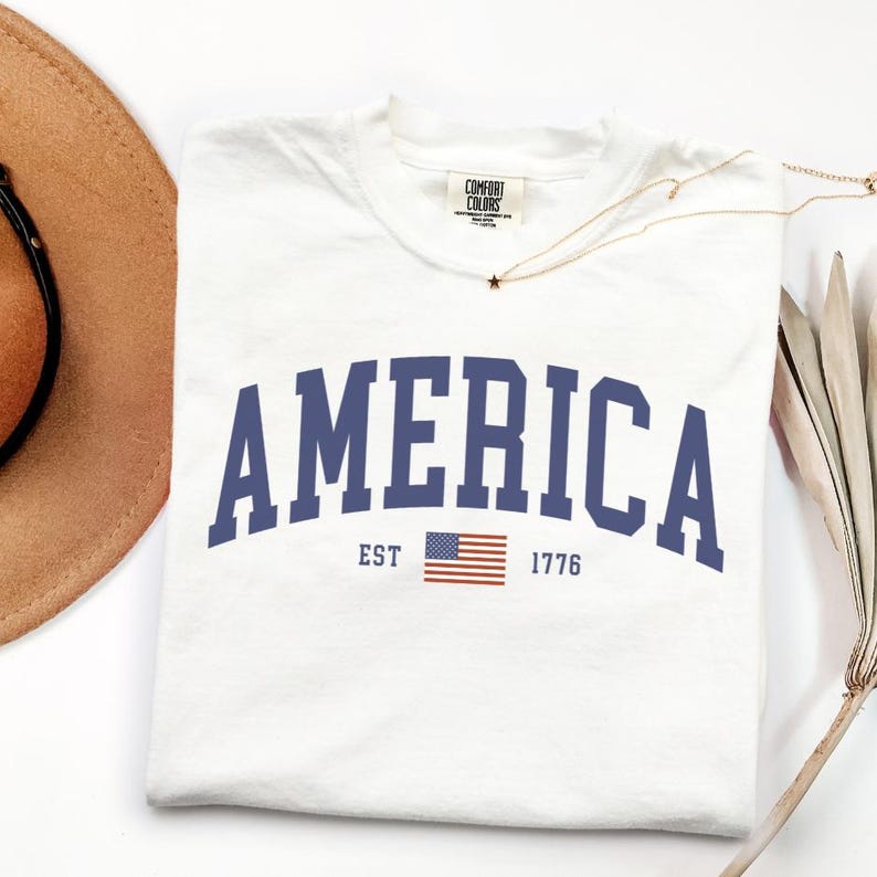 Peut inclure: T-shirt blanc avec le mot "AMERICA" en bleu marine, arqu&eacute; sur le devant. En dessous du mot "AMERICA" se trouve le texte "EST 1776" et un petit drapeau am&eacute;ricain. Le t-shirt a une &eacute;tiquette Comfort Colors.