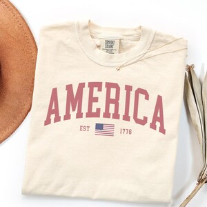 Peut inclure: T-shirt cr&egrave;me avec le mot "AMERICA" en gras, en rouge. En dessous, un petit drapeau am&eacute;ricain et le texte "EST 1776". Le t-shirt est de la marque Comfort Colors.
