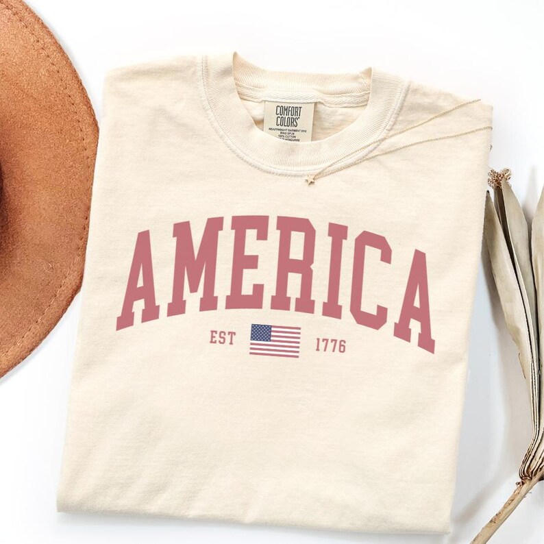 Peut inclure: T-shirt cr&egrave;me avec le mot "AMERICA" en gras, en rouge. En dessous, un petit drapeau am&eacute;ricain et le texte "EST 1776". Le t-shirt est de la marque Comfort Colors.