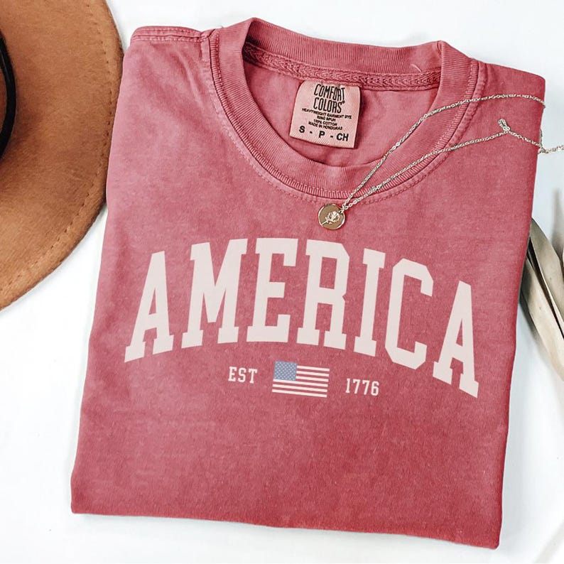 Peut inclure: Un t-shirt rouge avec le mot "AMERICA" en grandes lettres blanches. En dessous du mot "AMERICA" se trouve le texte "EST 1776" et un petit drapeau am&eacute;ricain. Un collier en or avec un pendentif est sur le t-shirt.