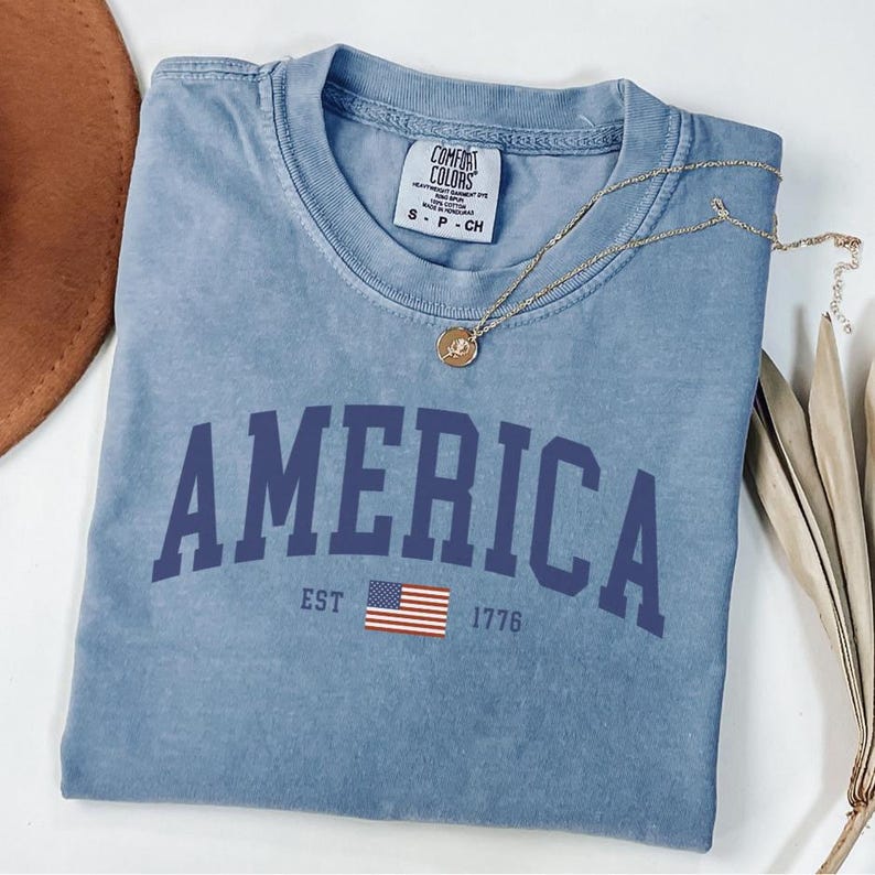 Peut inclure: Un t-shirt bleu clair avec le mot "AMERICA" en lettres majuscules bleu marine. En dessous, le texte "EST 1776" et un petit drapeau am&eacute;ricain. Un collier en or avec un pendentif repose sur le t-shirt.