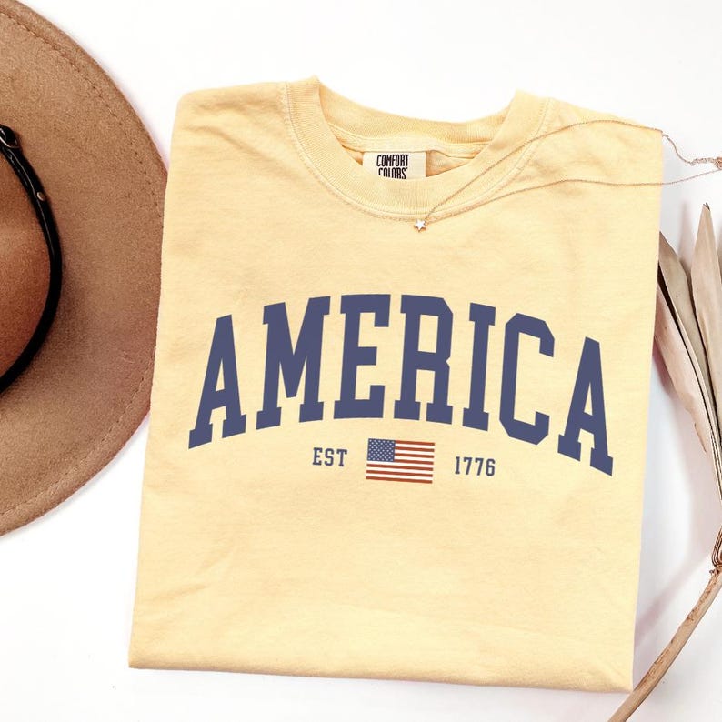 Peut inclure: Un t-shirt jaune clair avec le mot "AMERICA" en lettres majuscules bleu marine. En dessous, le texte "EST 1776" et un petit drapeau am&eacute;ricain. Le t-shirt est pli&eacute; et porte une &eacute;tiquette "Comfort Colors".