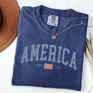 Peut inclure: Un t-shirt bleu marine avec le mot "AMERICA" en lettres majuscules bleu clair. En dessous, le texte "EST 1776" et un petit drapeau am&eacute;ricain. Un collier dor&eacute; avec un pendentif est pos&eacute; sur le t-shirt.