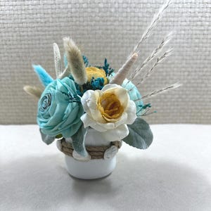 May include: A small, white ceramic vase with a nautical theme, adorned with rope and seashells. The vase holds a decorative arrangement of light blue, yellow, and white flowers, along with dried grasses and accents. The arrangement evokes a coastal or beach theme.