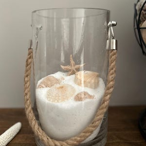 May include: A clear glass jar with a rope handle filled with white sand and seashells. The jar is decorated with a starfish and seashells.