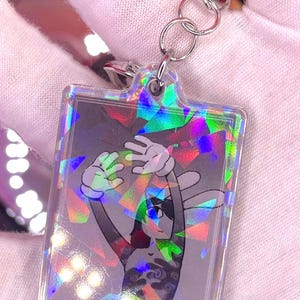 Peut inclure: Un porte-clés rectangulaire en acrylique avec une finition holographique à motif de verre brisé. Le design représente un personnage stylisé aux longues oreilles avec des gants blancs levés au-dessus de la tête, en nuances de gris et noir.