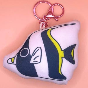 May include: A white, black, and yellow fish-shaped keychain. The fish has a dark blue stripe and yellow accents. The keychain has a pink metal clasp and ring, attached to the fish with a white cord. The background is a solid light pink.