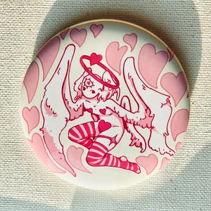 May include: A circular pinback button featuring a white background with a central illustration of a winged figure with a heart-shaped halo. The figure wears striped stockings and is surrounded by numerous pink hearts of varying sizes and shapes.