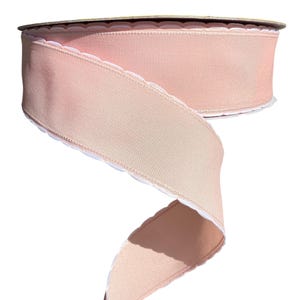 May include: A spool of blush pink ribbon with a textured weave. The ribbon has a scalloped white edge. The ribbon is partially unrolled, revealing the underside, which is a lighter shade of pink. This ribbon is suitable for crafts and gift wrapping.