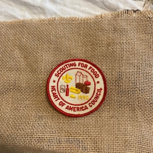 Puede incluir: Un parche circular con el texto "Scouting for Food" y "Heart of America Council" en rojo y blanco. El parche presenta ilustraciones de alimentos y el año 1990. El parche está sobre un fondo de arpillera.