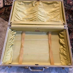 Puede incluir: Una maleta vintage abierta con un forro interior de tela de color amarillo claro. El interior cuenta con bolsillos laterales de tela fruncida, dos correas elásticas verticales para asegurar objetos y una pequeña etiqueta de marca rectangular.
