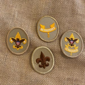 Puede incluir: Cuatro parches bordados con bordes beige y varios diseños. Dos parches presentan un emblema de águila y escudo, uno dice "BE PREPARED" en amarillo, y el último tiene un símbolo de flor de lis. Los parches están sobre una superficie de arpillera.