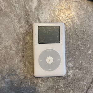 Könnte beinhalten: Ein weißer tragbarer digitaler Musikplayer mit einem rechteckigen Bildschirm und einem kreisförmigen Navigations-Clickwheel mit einer zentralen Auswahltaste. Das Gerät verfügt über eine Menütaste oben und Wiedergabesteuerungen für Titel.