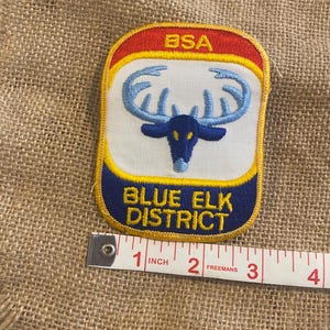 Puede incluir: Parche rectangular con borde superior rojo e inferior azul. El parche presenta un centro blanco con un diseño de alce azul y el texto "BLUE ELK DISTRICT". La parte superior del parche dice "BSA". El parche mide aproximadamente 5 cm de ancho.