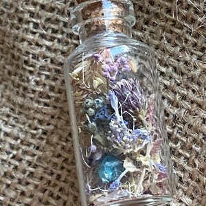 Op de afbeelding: Een klein glazen flesje met een kurken stop, gevuld met gedroogde bloemen en een blauwe edelsteen. Het flesje hangt aan een zilveren ketting.
