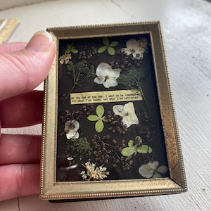 May include: A rectangular gold-toned metal frame containing a dark background with pressed dried flowers, green leaves, and small brown berries. A small paper strip in the center features the printed text: At the end of the day, I want to be remembered for what I've lived, not what I've collected.