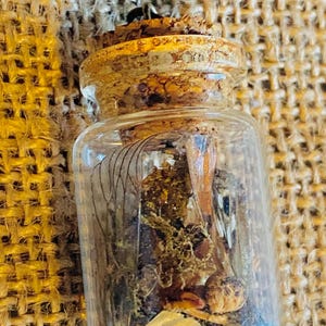 Puede incluir: Un pequeño frasco de vidrio con tapa de corcho lleno de botánicos secos y plumas. El frasco es transparente y el contenido es visible. El frasco está sentado sobre una superficie de arpillera.