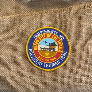 Könnte beinhalten: Ein kreisförmiger Aufnäher mit dem Text "Independence, MO" und "Queen City of the Trails" in einem blau-gelben Design. Der Aufnäher zeigt auch einen Planwagen und den Text "President Truman Trail" und "BSA 20 YRS".