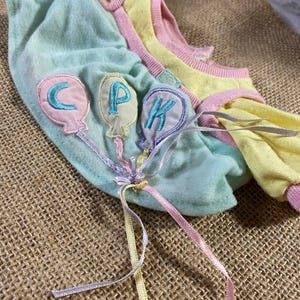 May include: A light blue and yellow doll dress with pink trim. The dress has three embroidered balloons with the letters CPK on them. The dress has three long ribbons attached to the balloons.