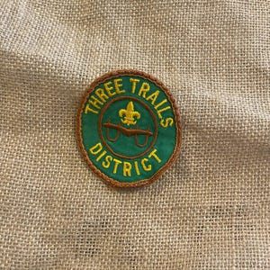 Peut inclure: Un écusson brodé vert et marron circulaire avec le texte "THREE TRAILS DISTRICT". L'écusson présente un motif de fleur de lis et de lunettes dorées. L'écusson est sur un fond de toile de jute beige.