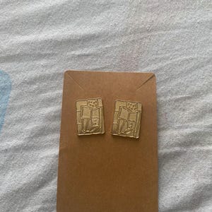Op de afbeelding: Twee goudkleurige vierkante oorstekers met een textuurontwerp, gepresenteerd op een bruine kaart. De oorbellen hebben een rechthoekige vorm met een reliëfpatroon, dat waarschijnlijk een gebouw of architecturaal element voorstelt.
