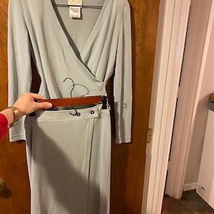 May include: A pale blue two-piece outfit, comprising a long-sleeved wrap top and a matching skirt. The top has a V-neck and the skirt has a button closure. The outfit is hanging on a wooden hanger.