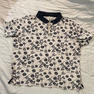 May include: A white polo shirt with a navy blue collar and short sleeves. The shirt is covered in a repeating pattern of dark blue floral vines and flowers. The shirt has a three-button placket.