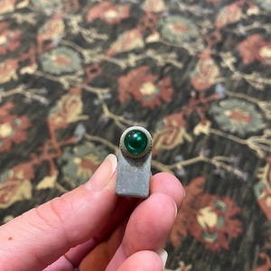 May include: A small, rectangular metal component featuring a circular, translucent green glass indicator light set into a textured, silver-toned metallic housing. The object has a flat base and a rounded top section that holds the green lens.