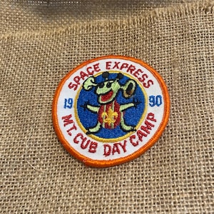 Pode incluir: Um patch circular com o texto "SPACE EXPRESS" e "MT. CUB DAY CAMP" em letras vermelhas. O patch apresenta um personagem de desenho animado de cachorro com um lenço vermelho e um fundo azul. O ano "1990" também é exibido.