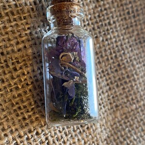 Op de afbeelding: Een klein glazen flesje met een kurken stop, gevuld met gedroogde bloemen en groen. Het flesje staat op een bruine jute achtergrond.