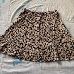 May include: A-line skirt with a floral pattern. The skirt features a navy blue background with a repeating pattern of small pink and cream-colored flowers. It has a button-up front closure.