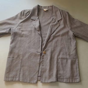 Könnte beinhalten: Ein hellbrauner Blazer mit einem dezenten Webmuster. Der Blazer hat einen Reverskragen, zwei Vordertaschen und einen Einzelknopfverschluss. Die Ärmel sind lang und das Kleidungsstück liegt flach.
