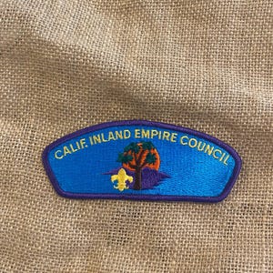 Puede incluir: Un parche azul y morado con el texto "CALIF. INLAND EMPIRE COUNCIL" en amarillo. El parche presenta un diseño de árbol, sol y flor de lis.