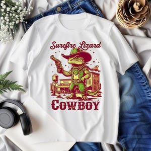 Puede incluir: Camiseta blanca con un lagarto de dibujos animados con un sombrero de vaquero y sosteniendo una pistola. El texto "Surefire Lizard Cowboy" está impreso en la camiseta.