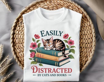 May include: White t-shirt with the text "EASILY DISTRACTED - BY CATS AND BOOKS." The shirt features a sleeping tabby cat on a stack of books, surrounded by pink flowers and green leaves. The design is printed on a white t-shirt.