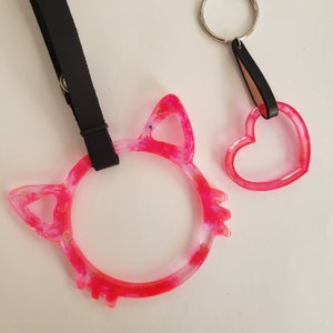 May include: Two pink resin keychains, one shaped like a cat and the other like a heart. Both keychains have a sparkly, translucent finish.