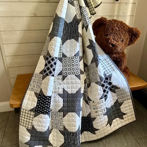 May include: A handmade quilt with a star pattern in shades of white, gray, and navy blue. The quilt is draped over a wooden bench, with a brown teddy bear peeking out from behind it. The quilt features various fabric patterns.