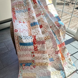 May include: A patchwork quilt with a variety of floral and geometric patterns in shades of red, blue, white, and beige. The quilt is folded and draped over a wicker chair.