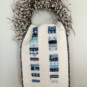 May include: A white quilted wall hanging with two vertical columns of blue fabric squares. The squares feature various patterns, including floral, plaid, and nautical designs. The hanging is framed by a navy blue border with white floral accents and is suspended from a white berry wreath.