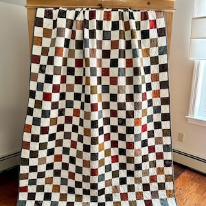 May include: A patchwork quilt featuring a grid of alternating white squares and various colored squares in shades of red, brown, green, and black. The fabric squares display diverse small-scale patterns and textures, all finished with a dark gray border.