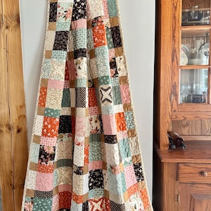 May include: A patchwork quilt with a variety of patterned squares in shades of pink, orange, brown, and green. The quilt is hanging from a metal clip and has a beige border. The quilt is likely handmade and has a vintage aesthetic.