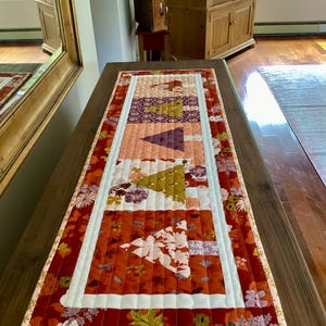 May include: A rectangular quilted table runner with a floral and geometric pattern. The runner features a red background with white and floral accents, and a central design of triangles and stripes in shades of orange, purple, and green. The runner is placed on a wooden table.