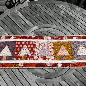 May include: A rectangular quilted table runner with a repeating tree design. The runner features a red floral border, white inner border, and four central squares with tree-shaped patterns in various colors, including burgundy, gold, and white.