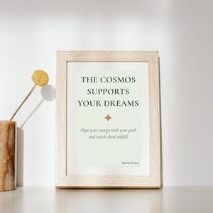 May include: A light wood frame containing a pale green print with the text THE COSMOS SUPPORTS YOUR DREAMS, a small four-pointed star icon, and the additional text Align your energy with your goals and watch them unfold and Words of The Stars.