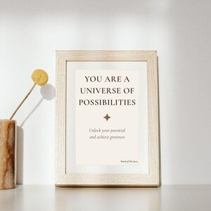 Può includere: Una cornice in legno chiaro contenente una stampa bianca con il testo YOU ARE A UNIVERSE OF POSSIBILITIES, una piccola icona a stella a quattro punte, il testo Unlock your potential and achieve greatness e Words of The Stars nell'angolo.