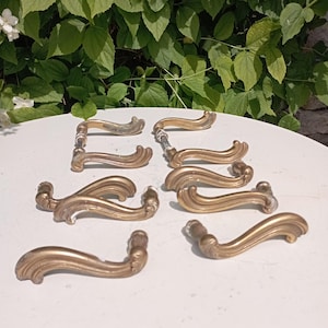 Pode incluir: Uma coleção de puxadores de porta antigos em latão com um design curvo e ornamentado. Os puxadores estão dispostos sobre uma superfície branca, com folhagem verde ao fundo. Cada puxador tem uma aparência detalhada e envelhecida.