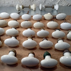Peut inclure: Une collection de boutons de porte anciens en porcelaine blanche avec des garnitures en laiton. Les boutons ovales sont disposés sur une surface en bois. Deux ensembles de boutons assortis sont en haut. Les boutons présentent de légères variations de couleur.