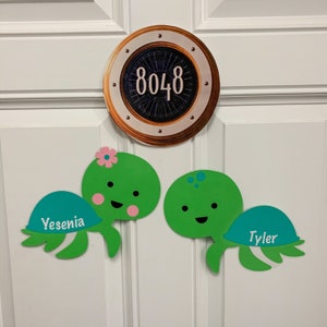 May include: A white door with a gold round porthole with the number 8048 on it. There are two green sea turtle decorations with names "Yesenia" and "Tyler" on them.